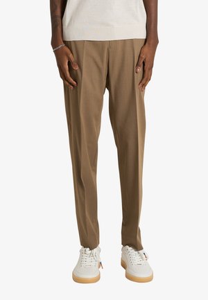 Parte inferiore del corpo di una persona che indossa pantaloni beige su misura, sneakers bianchi con suola in gomma e una camicia bianca a maniche corte, in piedi contro uno sfondo bianco.