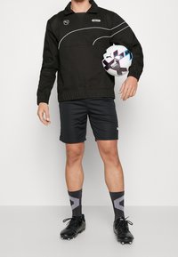 Giacca nera a pullover con dettagli bianchi, abbinata a pantaloni corti neri e scarpini neri. Tiene in mano un pallone da calcio con motivo, in piedi contro uno sfondo neutro.