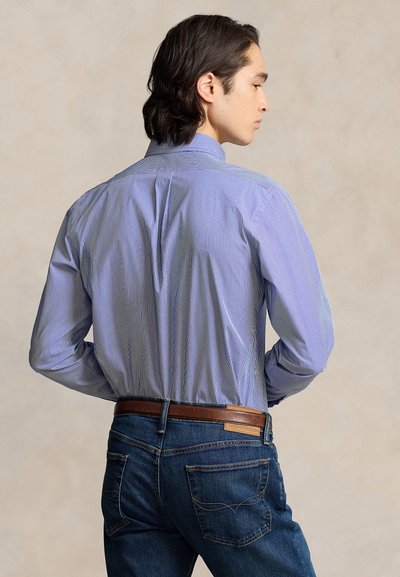 Fiatal férfi hosszú sötét hajjal, kék csíkos ingben, berakva kék farmernadrágba, barna bőr övvel, háttal nézve.