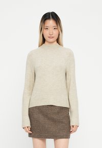 Beige neule, jossa on korkea kaulus, ribsikuvioitu helma ja pitkät hihat, yhdistettynä ruskeaan houndstooth-hameeseen, joka on muotoilultaan tiukka.