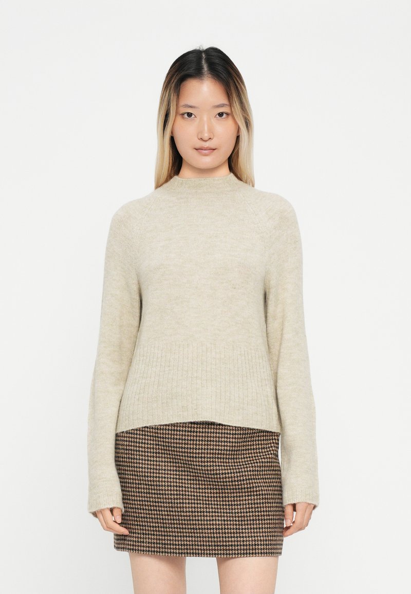 Beige neule, jossa on korkea kaulus, ribsikuvioitu helma ja pitkät hihat, yhdistettynä ruskeaan houndstooth-hameeseen, joka on muotoilultaan tiukka.