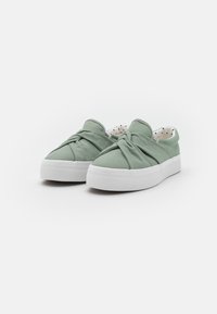 Zapatillas verdes de lona sin cordones con un diseño anudado en la parte superior y una suela de goma blanca. Forradas con tela de lunares por dentro.
