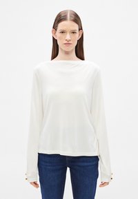 Hosszú ujjú krémszínű felső, sima anyagból, csónaknyakkivágással. Az ujjvégeken gombdíszítéssel és laza szabással.