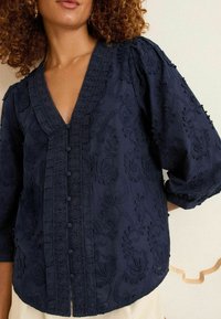 Blusa ricamata blu navy con scollatura a V, maniche a palloncino, intricati motivi floreali e bottoni lungo la parte anteriore. Tessuto testurizzato.