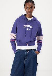 Hoodie cropped roxo com o logótipo branco "Converse All Star", detalhes nas mangas em rosa e lavanda, e cordão ajustável na barra. Usado com calças de ganga largas.