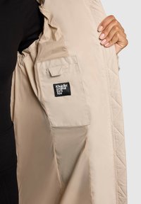 Interiorul jachetei bej, captusita, cu o eticheta, avand un buzunar lateral cu clapeta si o textura de material neted si lucios.