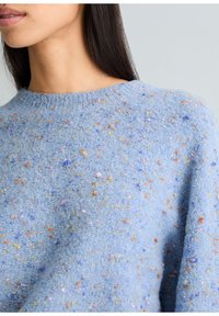 Femme portant un pull en tricot bleu clair avec des taches multicolores, montrant la zone du cou et des épaules avec des cheveux raides et foncés.