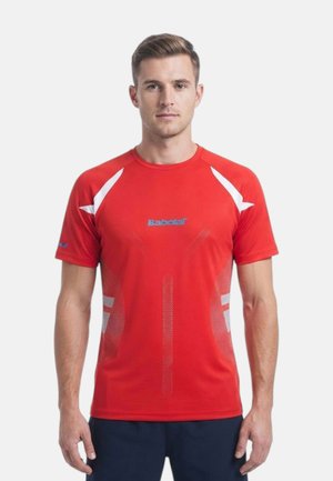 Uomo con capelli corti che indossa una maglietta sportiva rossa con dettagli bianchi e logo blu, in piedi davanti a uno sfondo chiaro e uniforme.