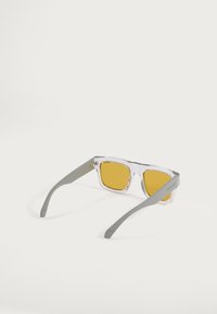 Gafas de sol rectangulares transparentes con lentes amarillos y patillas rectas grises, colocadas mirando hacia atrás sobre una superficie blanca.