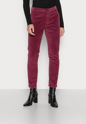 Žena má na sobě úzké vínové kalhoty z manšestru, černý top s dlouhým rukávem a černé kotníkové boty s blokovým podpatkem na jednoduchém světlém pozadí.