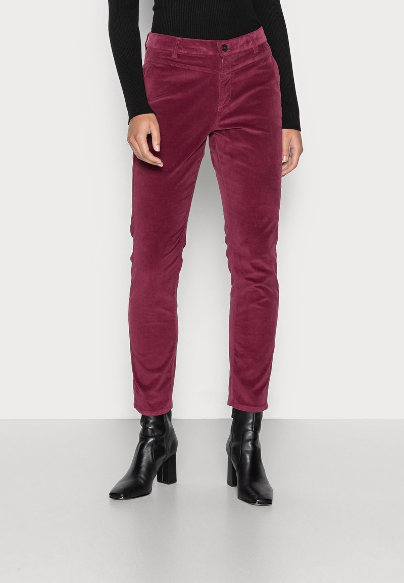 Femme portant un pantalon en velours côtelé bordeaux ajusté, un haut à manches longues noir et des bottines noires à talons blocs, sur un fond clair uni.