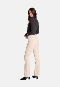 Modèle portant un pull côtelé sombre et un pantalon évasé beige clair, avec des bottes à talons noires. Le pull présente des poignets boutonnés.