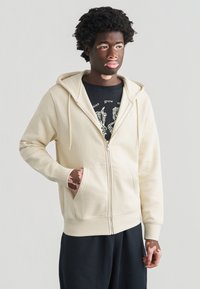 Ανοιχτό μπεζ φούτερ με φερμουάρ μπροστά, κουκούλα με κορδόνι και τσέπες μπροστά. Διαθέτει ριμπ μανσέτες και τελείωμα, φορεμένο πάνω από ένα μαύρο φανελάκι με στάμπα.