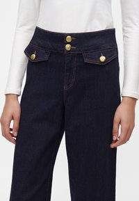 Jean bleu foncé taille haute avec jambes larges, deux poches avant à rabat et boutons décoratifs dorés, porté avec un haut blanc manches longues.