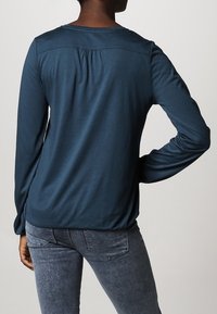 Langärmliges, tiefblaues Oberteil aus weichem Stoff, mit einem gerafften Detail auf der Rückseite, kombiniert mit enganliegenden grauen Jeans.