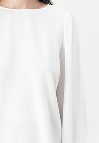 White long-sleeve top with a round neckline, featuring a textured fabric and soft drape, highlighting gentle volume in the sleeves.