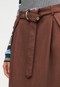 Καφέ παντελόνι με ψηλή μέση και ζώνη από το ίδιο ύφασμα με αγκράφα D-ring από χρυσό μέταλλο, φορεμένο με πολύχρωμο πλεκτό τοπ με εμφανές μανίκι.