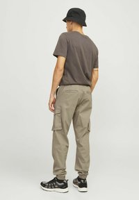 Mann trägt einen schwarzen Fischerhut, dunkelgraues T-Shirt, beige Cargo-Hosen und schwarze Sneaker, steht mit dem Rücken zur Kamera vor einem einfarbigen Hintergrund.