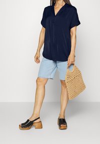 Camicia navy a maniche corte, pantaloni corti in denim chiaro, zoccoli neri con tacco in legno e una borsa intrecciata beige. Outfit casual con vestibilità rilassata e texture.