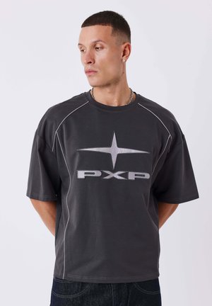 Uomo con capelli corti e scuri che indossa una maglietta scura oversize con stella e logo PXP, guarda verso sinistra su uno sfondo chiaro uniforme.