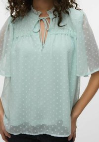 Ljus mintgrön blus med prickigt mönster, rynkad halsringning, knytband och korta, genomskinliga ärmar. Diskret spetskant framtill.