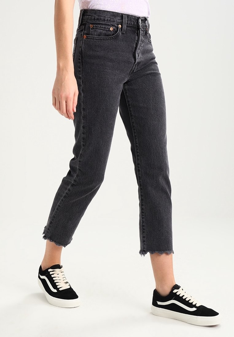 Personne portant un jean noir taille haute court avec des ourlets bruts et des baskets noir et blanc, debout sur un fond blanc.