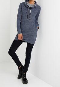 Abito felpa blu navy con un ampio colletto grigio e lacci a contrasto, abbinato a jeans skinny neri e stivali neri con lacci.