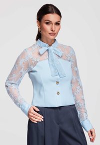 Femme portant un chemisier bleu clair avec des manches en dentelle et un col à nœud papillon, associé à un pantalon sur mesure bleu marine foncé.