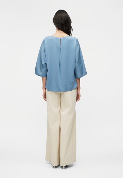 Γυναίκα με μακριά σκούρα μαλλιά φοράει ένα άνετο ανοιχτό μπλε τοπ με μανίκια μέχρι τον αγκώνα και εκρού παντελόνια με φαρδιά μπατζάκια, στέκεται μπροστά σε ένα απλό φόντο.