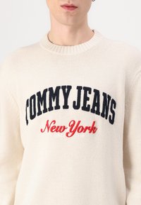 Pulover tricotat crem, cu broderie în negru "TOMMY JEANS" și "New York" în roșu, având un guler rotund ribanat și un material texturat.