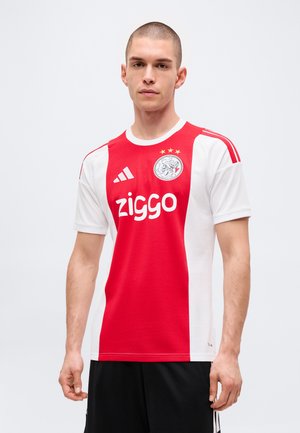 Młody mężczyzna z krótkimi włosami, ubrany w czerwono-biały strój piłkarski Ajax z logo sponsora Ziggo i Adidas, stojący na jednolitym jasnym tle.