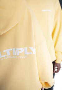 Sudaderas amarillas de tejido suave con un logo blanco de "MULTIPLY APPAREL CLUB" en el pecho, un corte relajado y una capucha grande.