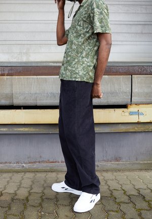 Mężczyzna w zielonej, kwiecistej koszuli, czarnych spodniach i białych tenisówkach stoi na tle przemysłowej metalowej ściany, trzymając czarny pasek blisko twarzy.