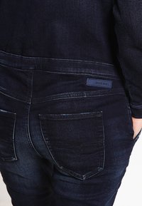 Gros plan d'un jean Diesel bleu foncé montrant la poche arrière et le patch logo sur la ceinture, avec la main d'une personne partiellement dans la poche avant.