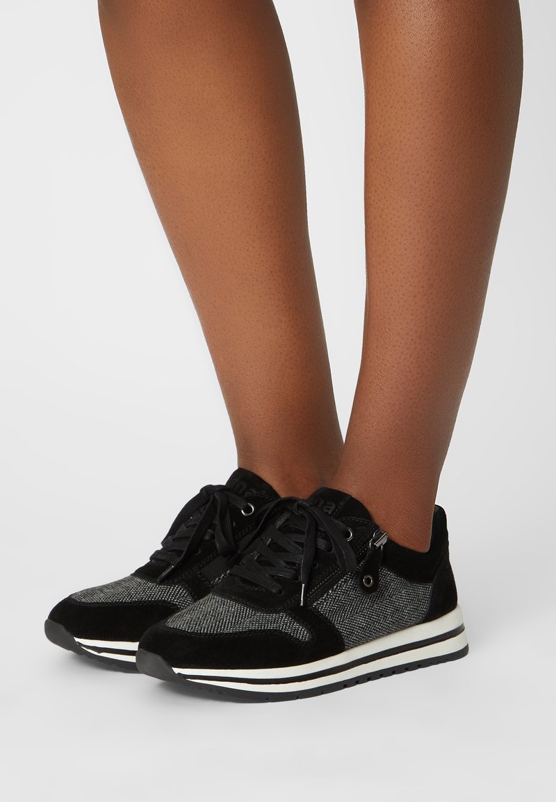 Baskets basses noires et grises avec lacets et fermeture éclair latérale portées sur les jambes à la peau foncée d'une personne sur fond blanc.