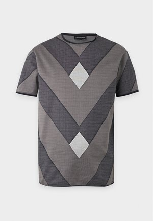 Mand stående med hænderne i lommerne iført en grå T-shirt med geometrisk mønster, sorte bukser, bælte og sorte loafers mod en ensfarvet baggrund.
