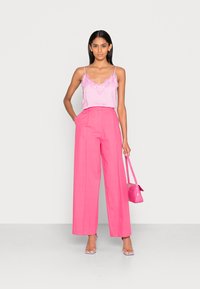 Camisola de cetim rosa claro com bordado em renda, combinada com calças largas de cintura alta rosas. Modelo segura uma bolsa rosa combinando.