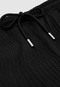 Tissu noir côtelé avec une ceinture élastique et un cordon noir noué avec des embouts en métal argenté.