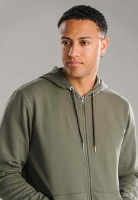 Zipper hoodie i olivengrøn, lavet af blødt stof. Har en hætte med snoretræk, to forlommer og sølvfarvede hardware-detaljer.