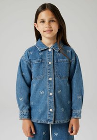 Džinsa jaka ar pogu priekšējo daļu, divām krūšu kabatām, izšūtiem lokiem un klasisku apkakli. Krāsa ir vidēji zila ar nelielu izbalējumu.