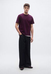 Maroon cotton t-shirt with a subtle logo, paired with loose-fitting dark denim jeans. Model stands against a neutral backdrop.