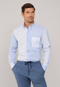 Camicia button-up con righe verticali blu e bianche, colletto e taschino. Indossata con pantaloni blu a vestibilità rilassata, dotati di cordoncino.