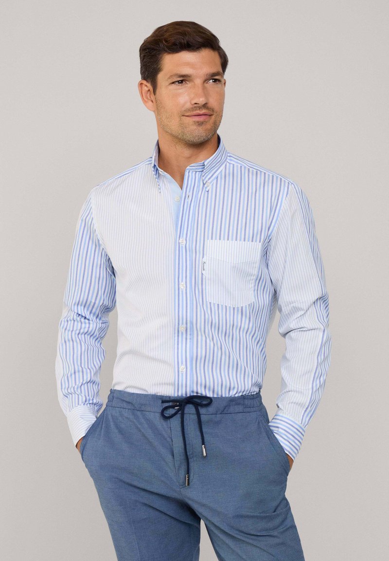 Camicia button-up con righe verticali blu e bianche, colletto e taschino. Indossata con pantaloni blu a vestibilità rilassata, dotati di cordoncino.