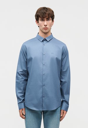 Vyras dėvintis paprastą mėlyną ilgomis rankovėmis marškinius su sagomis ir šviesiai mėlynas džinsas, stovintis neutraliame fone.