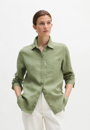 Kobieta w jasnozielonej, zapinanej na guziki koszuli z długim rękawem i przednią kieszenią oraz białych spodniach, stojąca z rękami w kieszeniach.