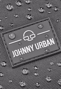 Tessuto grigio con texture e gocce d'acqua; un'etichetta di gomma rettangolare recita "JOHNNY URBAN" in bianco, con un'illustrazione di occhiali sopra il testo.