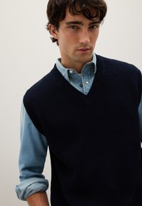 Un gilet in maglia blu navy con scollo a V indossato sopra una camicia di denim azzurro chiaro, che mostra un tessuto texturizzato e una vestibilità comoda.