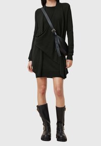 Femme portant une robe noire asymétrique à manches longues, un sac bandoulière noir et des bottes noires mi-mollet à boucles.