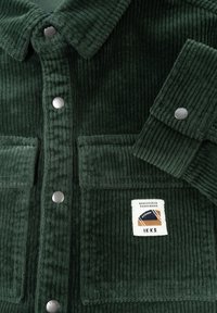 Giacca in velluto a coste verde con colletto, bottoni a pressione in argento, due tasche frontali e una patch con design geometrico.