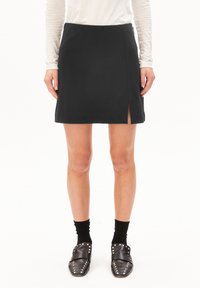 Frau, die einen schwarzen Rock mit Schlitz vorne, schwarze mit Nieten besetzte Slipper, schwarze Socken und ein helles Langarmoberteil trägt, steht vor weißem Hintergrund.